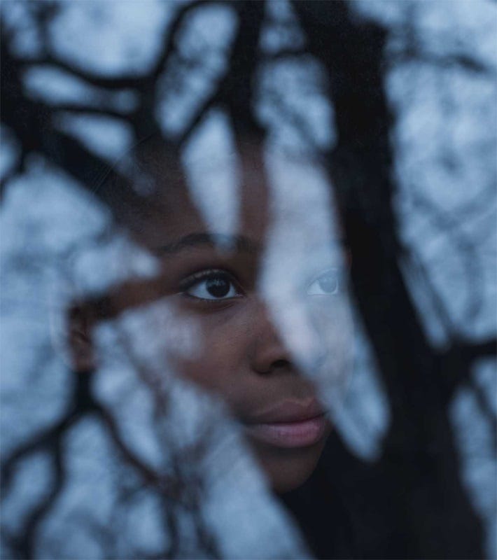 A color photograph of a young Black boy looking through a window, his image partly obscured by the reflection of bare trees and a wintry sky. He gazes into the sky, a neutral expression on his face that could be uncertainty or could be hope. 