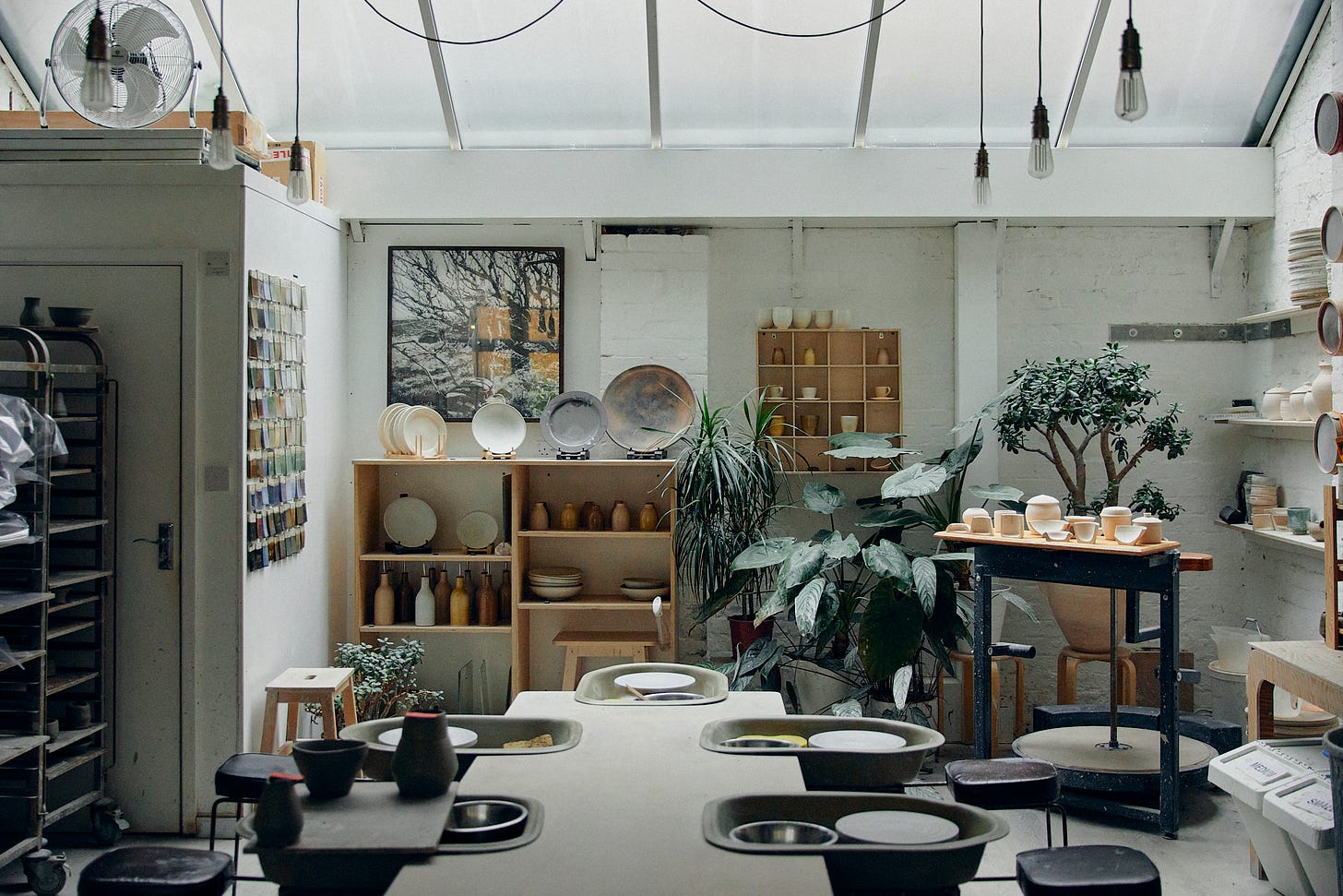 Inside a ceramics studio. A large table runs through the centre foreground with pottery wheels. Plants along the back wall, shelves on all sides with display objects; bowls, plates, vessels. A roof light at the top letting in soft light. A picture on the back wall of trees in winter.