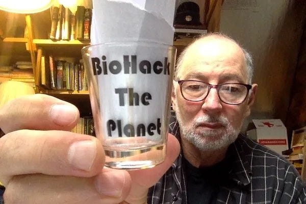 Photo of some old guy in a cluttered office holding to the camera a shot glass that has 'BioHack the Planet" written on it in bold type. There is a piece of white paper in the glass to make the printed words stand out. Photo of some old guy in a cluttered office holding to the camera a shot glass that has 'BioHack the Planet" written on it in bold type. There is a piece of white paper in the glass to make the printed words stand out.