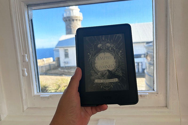 A hand holding a Kindle with a copy of ‘Empire of the Damned’ on it by Jay Kristoff. In the background is a light house.