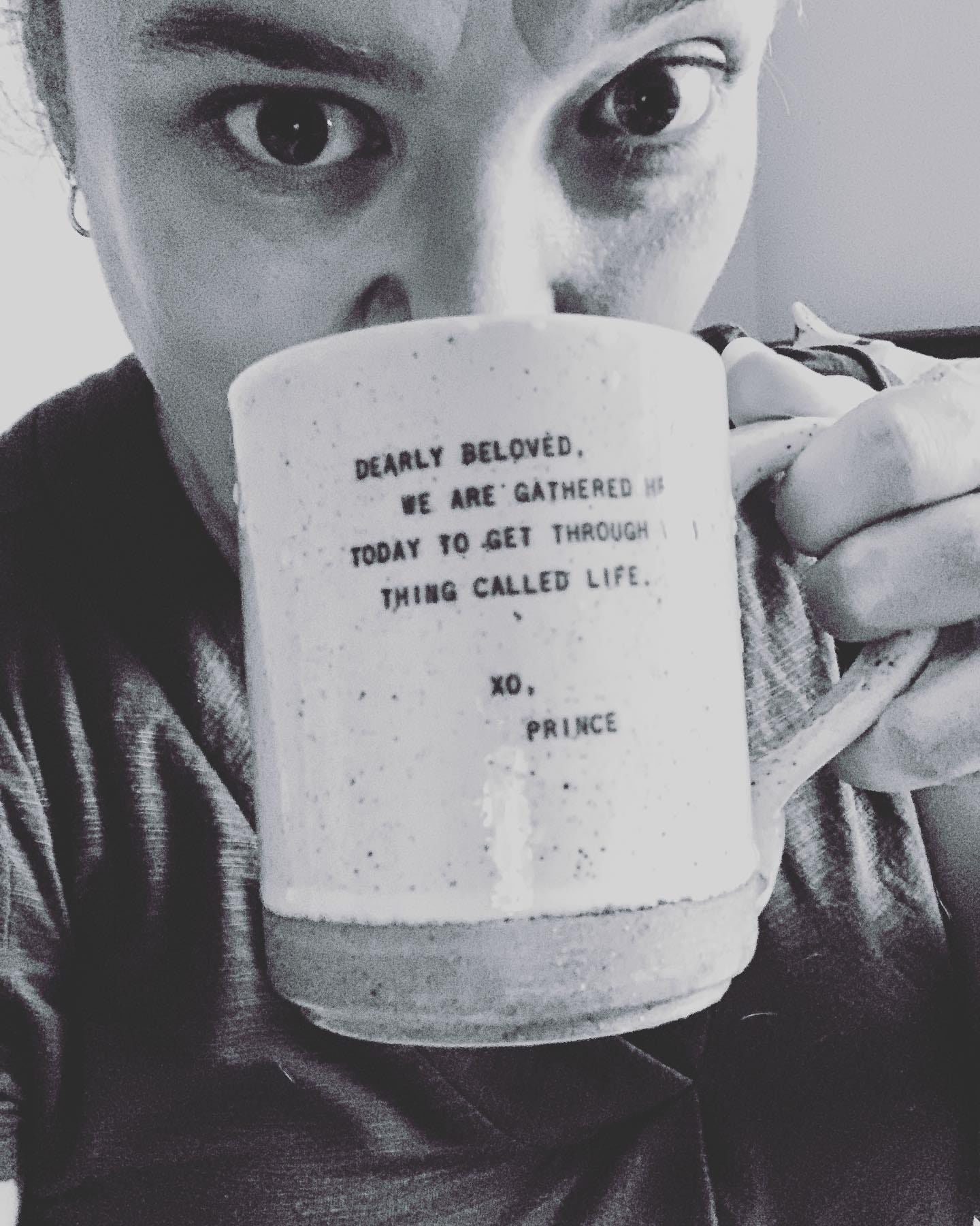 A woman with her hair in a bun and large brown eyes drinking out of a coffee mug that has the Prince lyrics "Dearly Beloved, we are gathered here today to get through this thing called life." printed on the side
