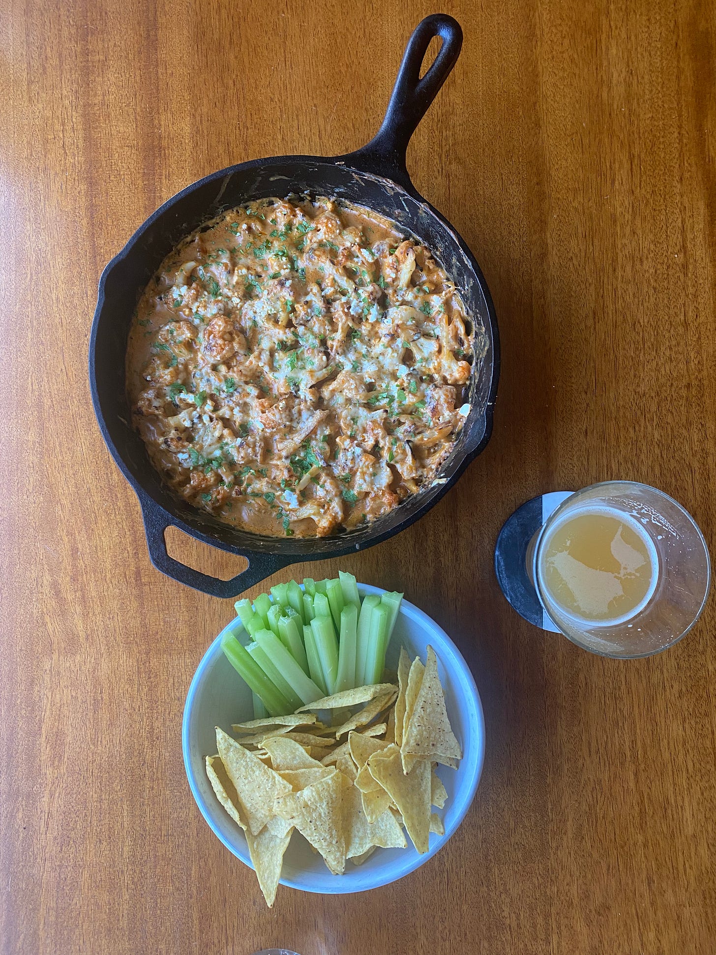 From above, a white bowl of yellow tortilla chips on one side and celery sticks on the other. Above the bowl is a cast-iron pan of the dip described above, creamy orange with cheese and parsley on top. A glass of beer sits on a coaster to the right.