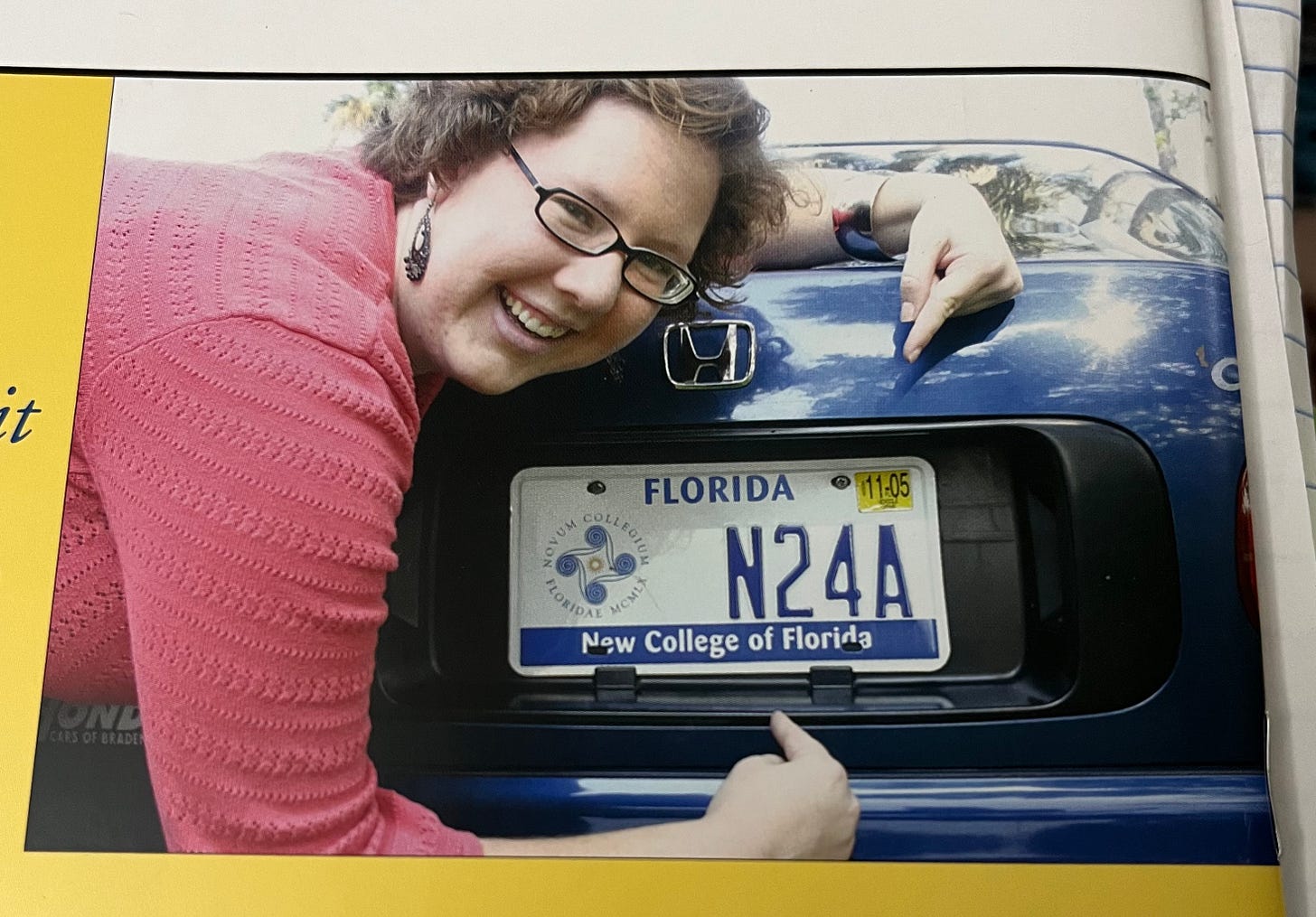 Photo of a white woman with curly reddish-hair and glasses pointing to a New College license plate as part of an advertisement