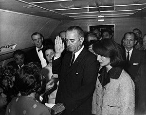 Lyndon B. Johnson raises his hand above an outstretched Bible as his is sworn in as President as Air Force One prepares to depart Love Field in Dallas. Jacqueline Kennedy, still in her blood-spattered clothes (not visible), looks on.