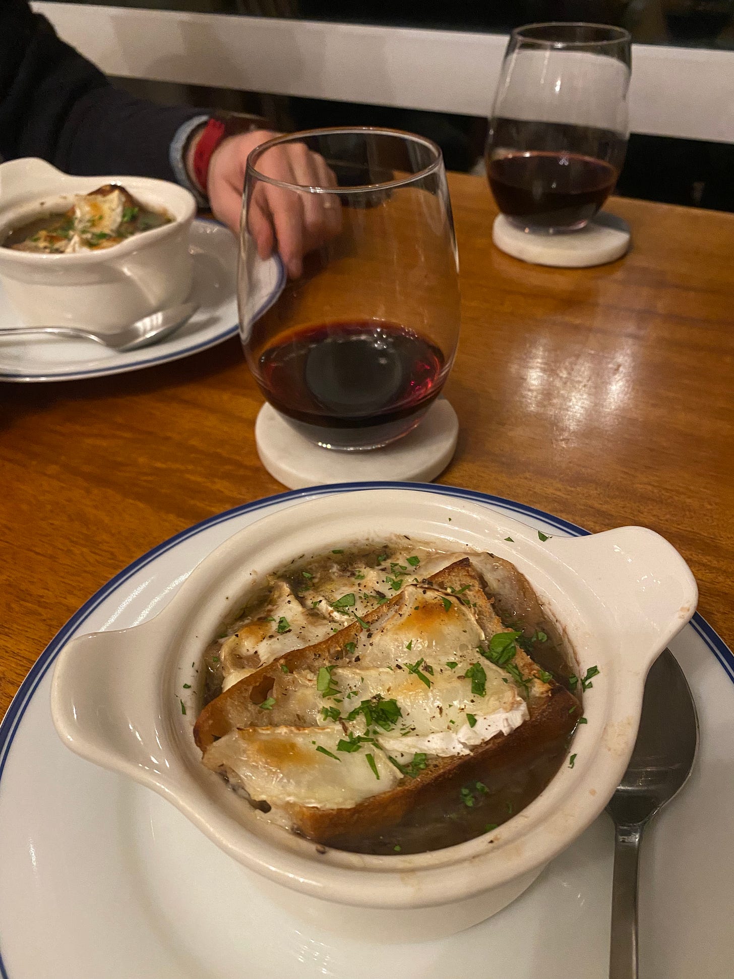 Two small plates with white french onion soup bowls on them, filled with rich brown soup and topped with slices of cheesy sourdough. Parsley and black pepper are scattered over the top. On coasters near the plates are glasses of red wine in stemless glasses.