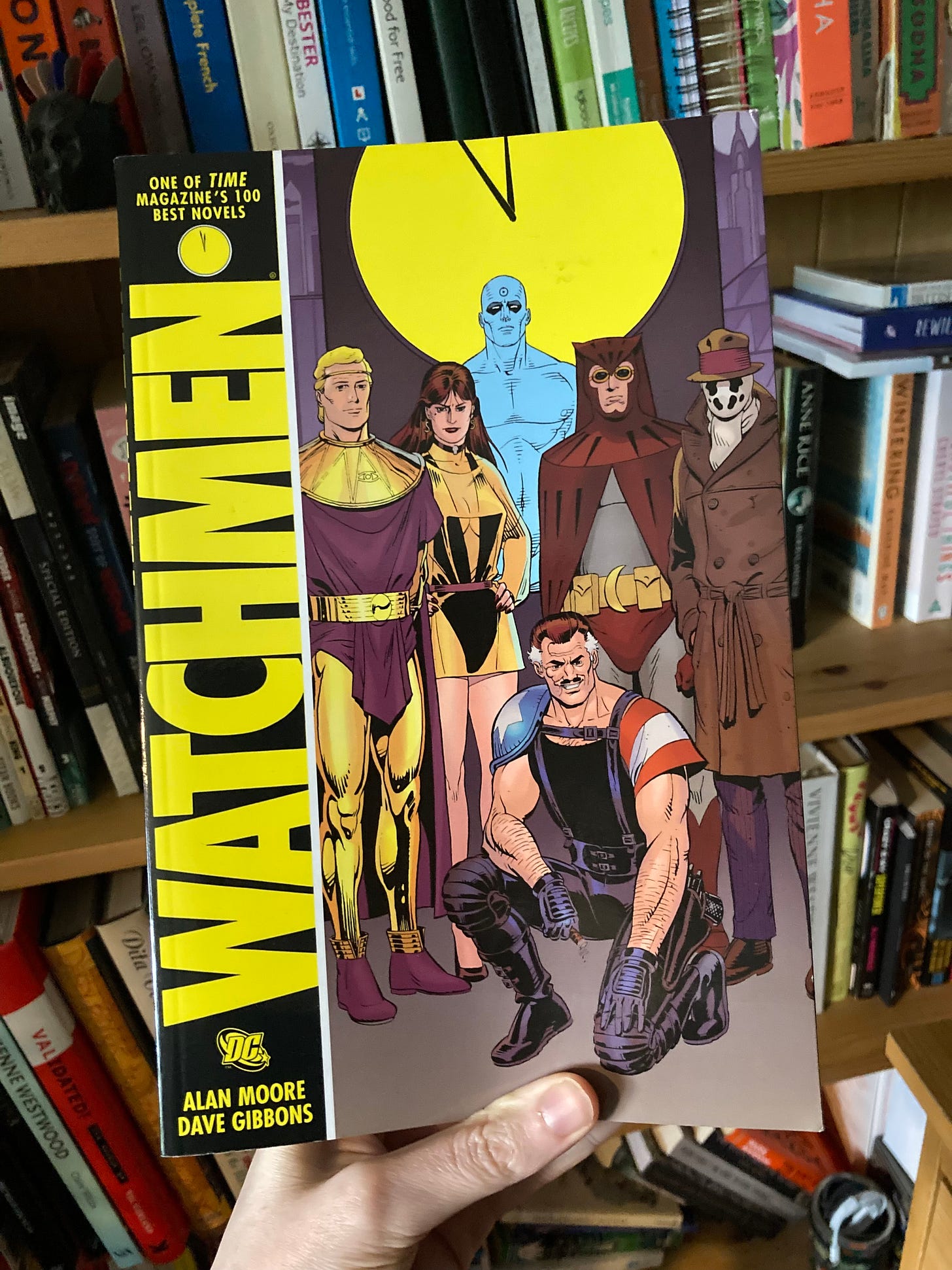 Watchmen written by Alan Moore and illustrated by Dave Gibbons being held in my hand, in front of my bookshelf