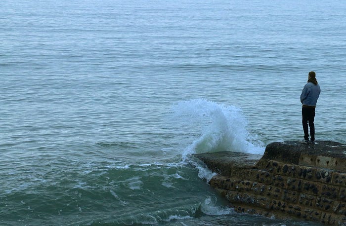 a person, probably a man, looking thoughtfully out over an outcropping onto the sea as a wave breaks