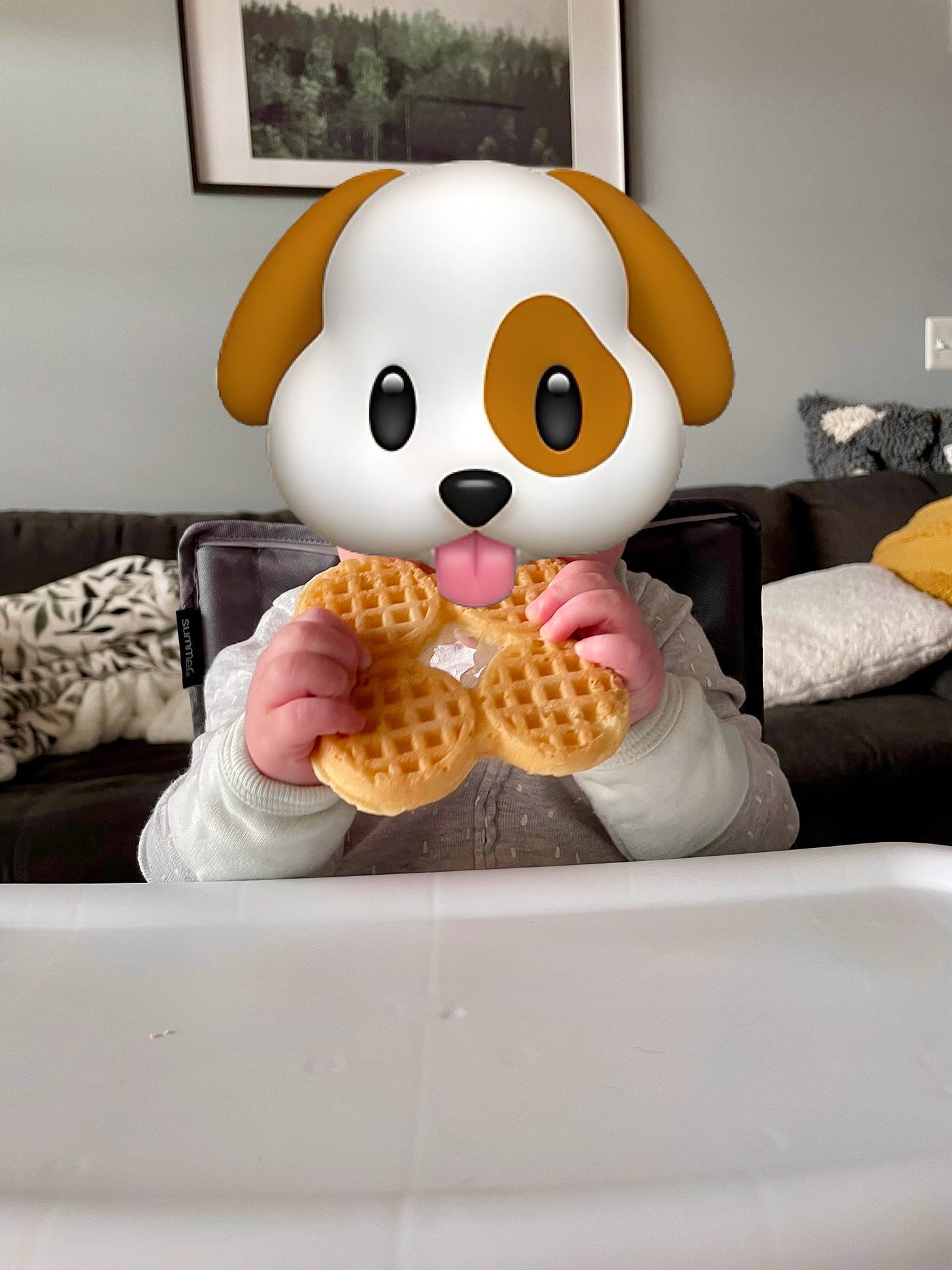 A baby sits in a high chair two fisting a waffle. A dog sticker covers her face. 