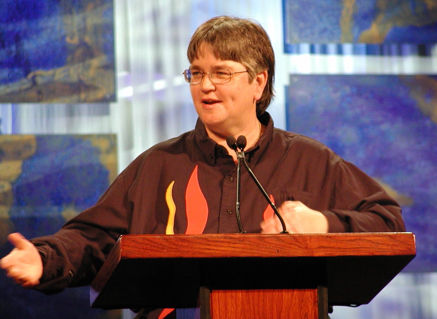 A woman with short brown hair and glasses wearing a shirt with flames on it standing at a podium