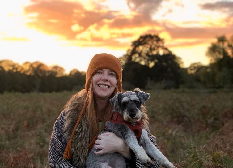 Allie is standing in a field, holding Dottie in her arms. Behind them is a line of trees and a beautiful sunset comprising orange, yellow, pinks and purples. 