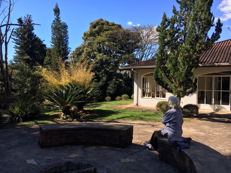 The entrance to a brazilian guesthouse, with a blue sky and various types of trees. An elderly woman sits with her back to the camera in front of a firepit. 