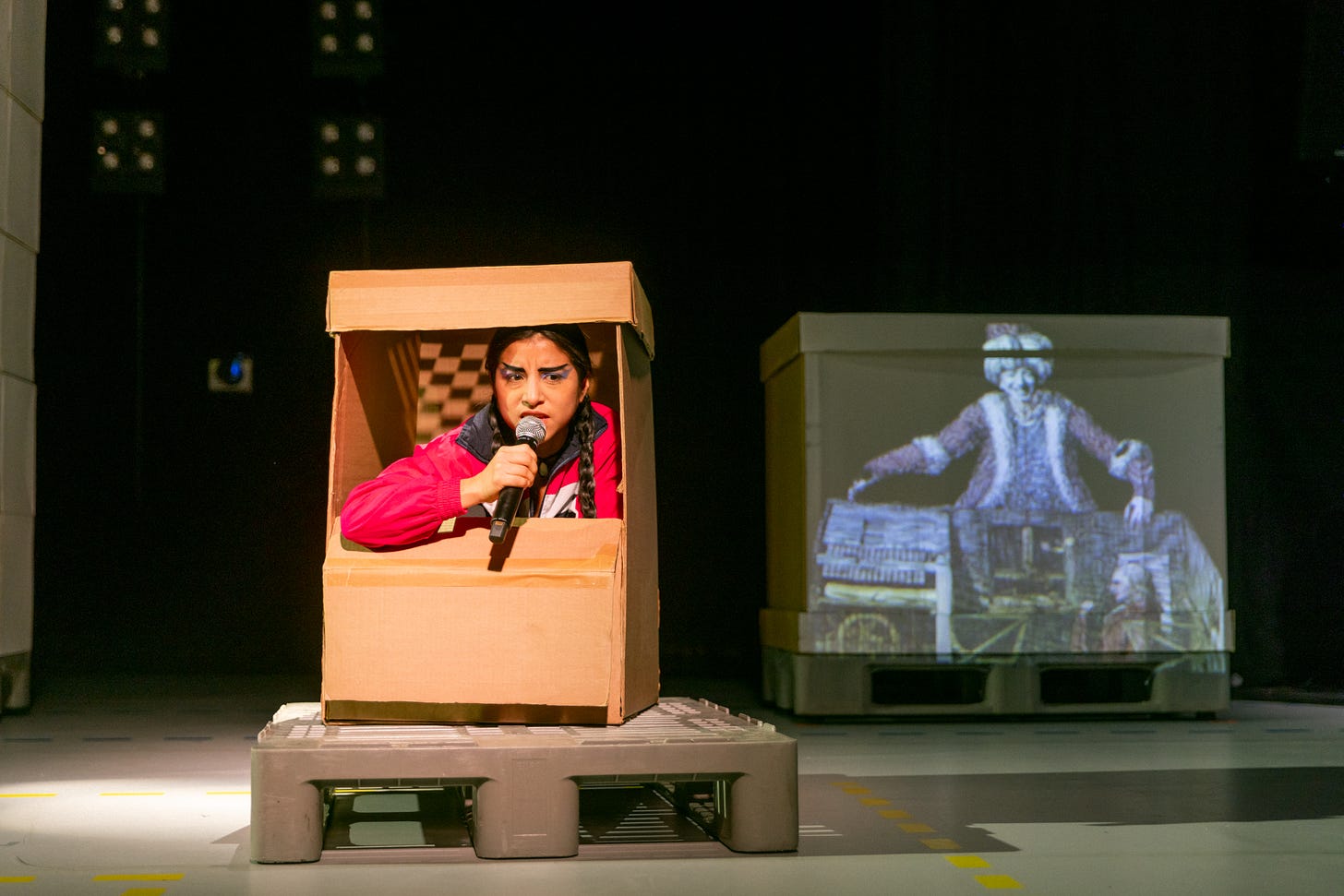 A circus performer sits inside a cardboard box holding a microphone.