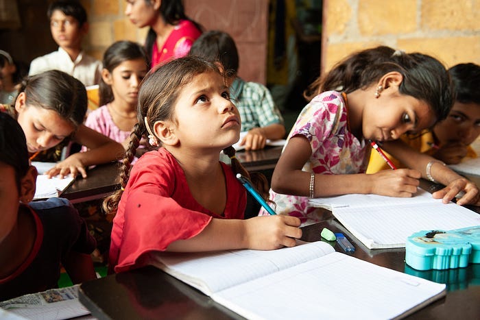 A little girl with a notebook and pencil looking up at the board in a classroom, amidst a whole bunch of other students