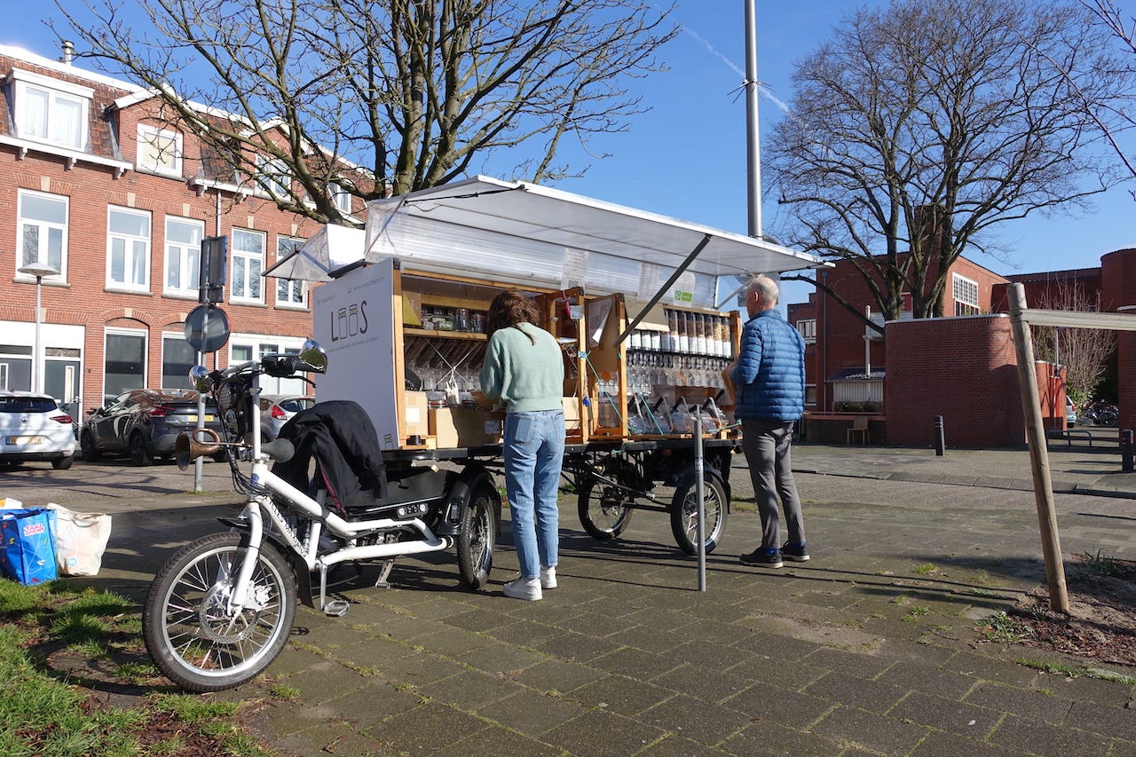A tricyle cargobike and its large trailer are parked on a public square in a residential neighbourhood. Connected to the back of the bike is a 3 meter long trailer. On top of the trailer is a roughly 1 meter high display of serving containers. Inside the containers are seeds, nuts, cereals etc. The woman who drives the bike and and owns the bike is preparing a customers order as he waits next to her. 