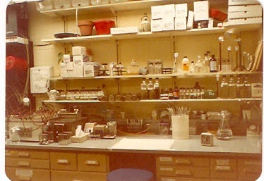 A yellowed photograph of a bench in a molecular biology lab. There is a desk with notebooks, and shelves full of various chemical and glassware. Everything is as neat as a pin.