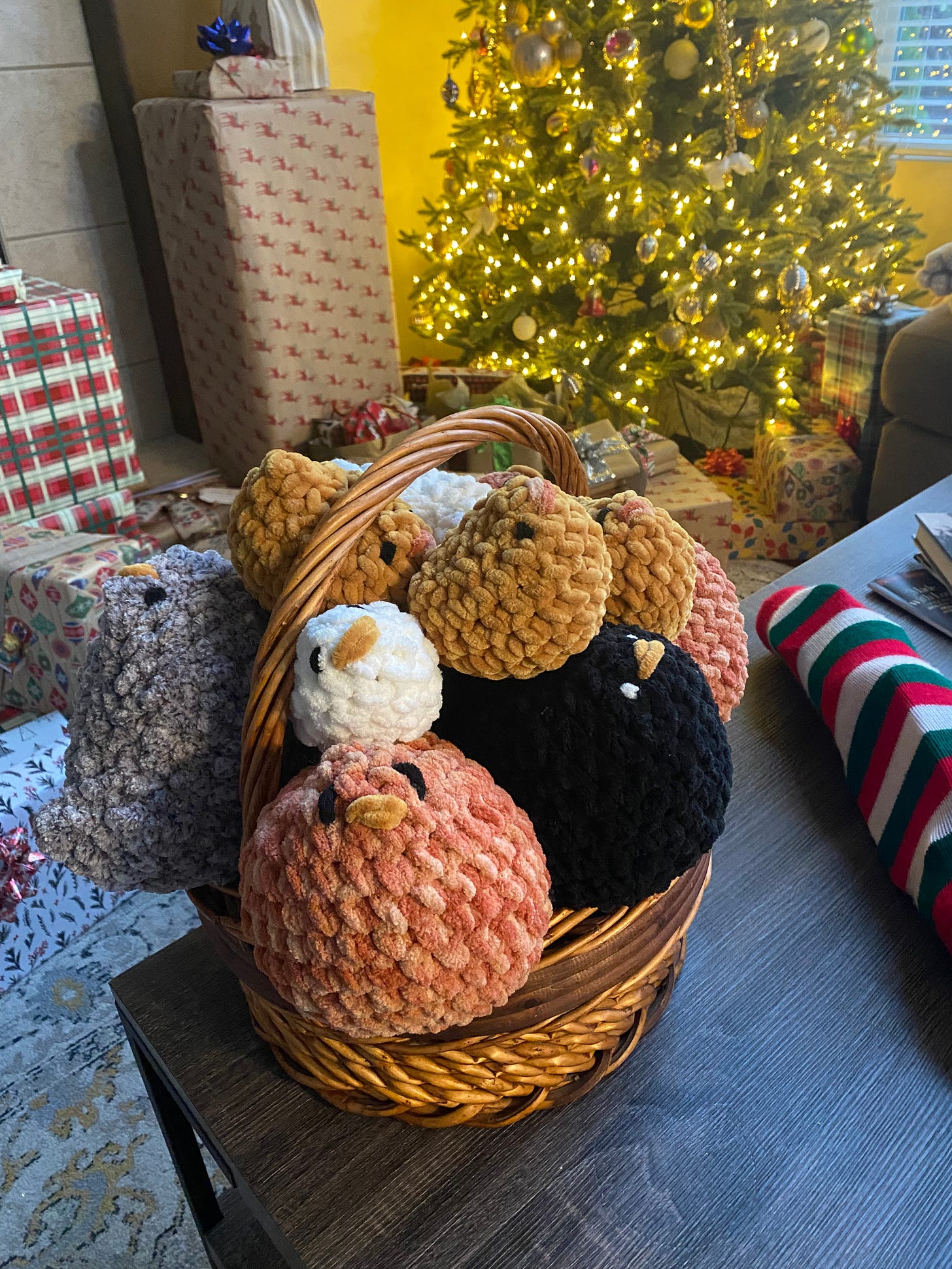 On a coffee table in front of a Christmas tree with wrapped gifts around it, a basket of puffy, crocheted chickens of various sizes and colours.