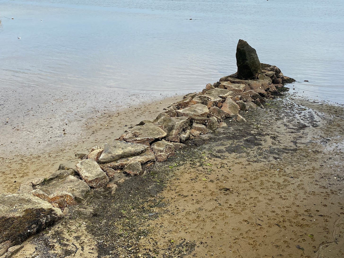 A photograph of a stone breakwater at low tide. The stones are piled into a barnacle-encrusted line ending in a tall and pointed monolith. Beyond the sand and stones are the still waters of a harbor.