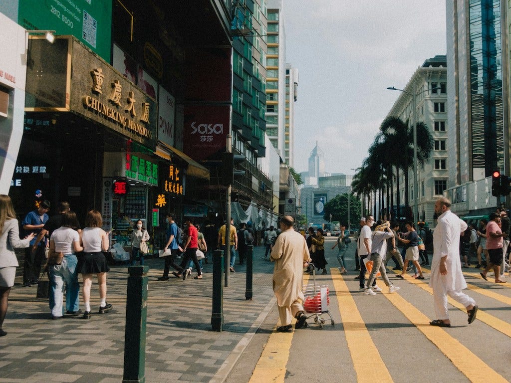 Foto a colori di una strada di Hong Kong. Si vedono diverse persone che camminano e attraversano la strada, e alcuni grattacieli. Sulla sinistra, c'è l'ingresso alle Chungking Mansions.