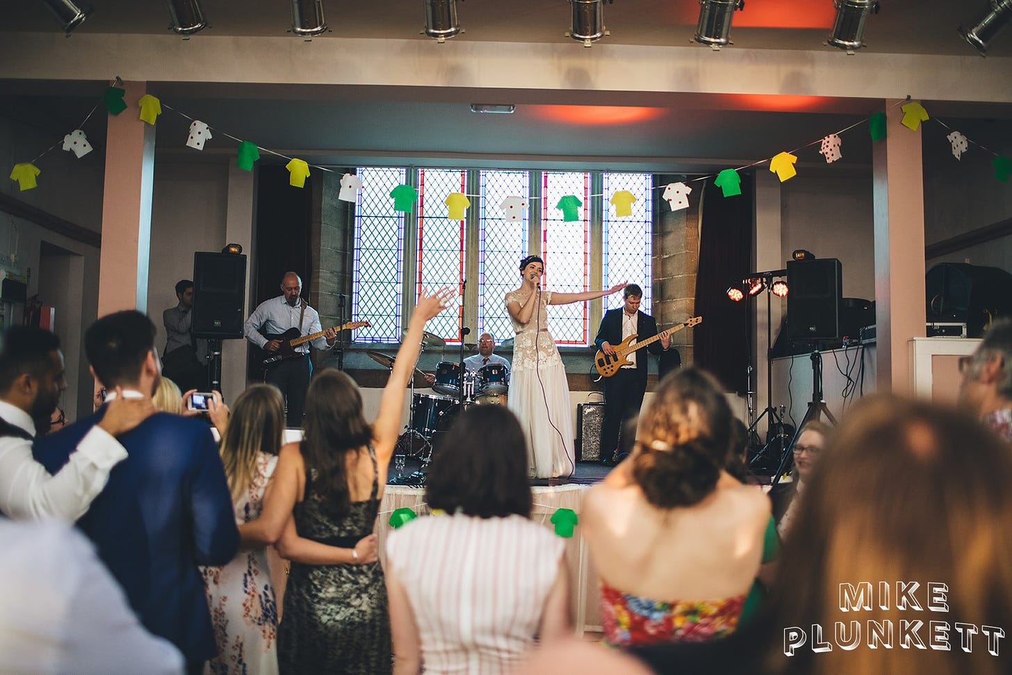 I’m a white woman with short brown hair, in the middle of the stage with my right arm out by my side wearing a beautiful tulle-skirt wedding gown. Backed my Jon on bass, Andy on guitar and Steve on drums. My loved ones are in the foreground watching from the floor.