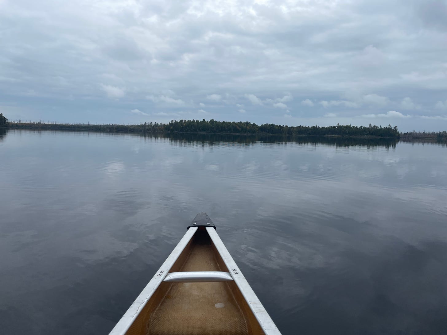 The bow of a canoe points across a still northern lake toward a distant shore of pine trees. The cloudy sky is reflected in the lake.