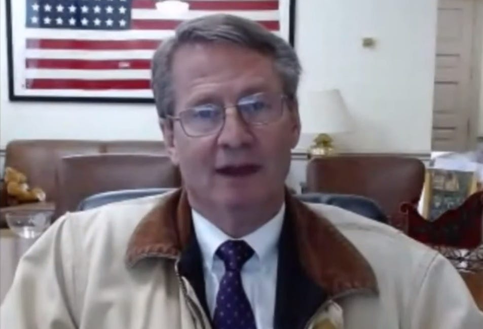 Screenshot of Rep Tim Burchett (R-Tennessee) on a livestream from his office, wearing a white shirt and tie but with a corduroy-collared casual coat. Behind him are a framed US flag with 48 stars and a couch on which sits a teddy bear. 