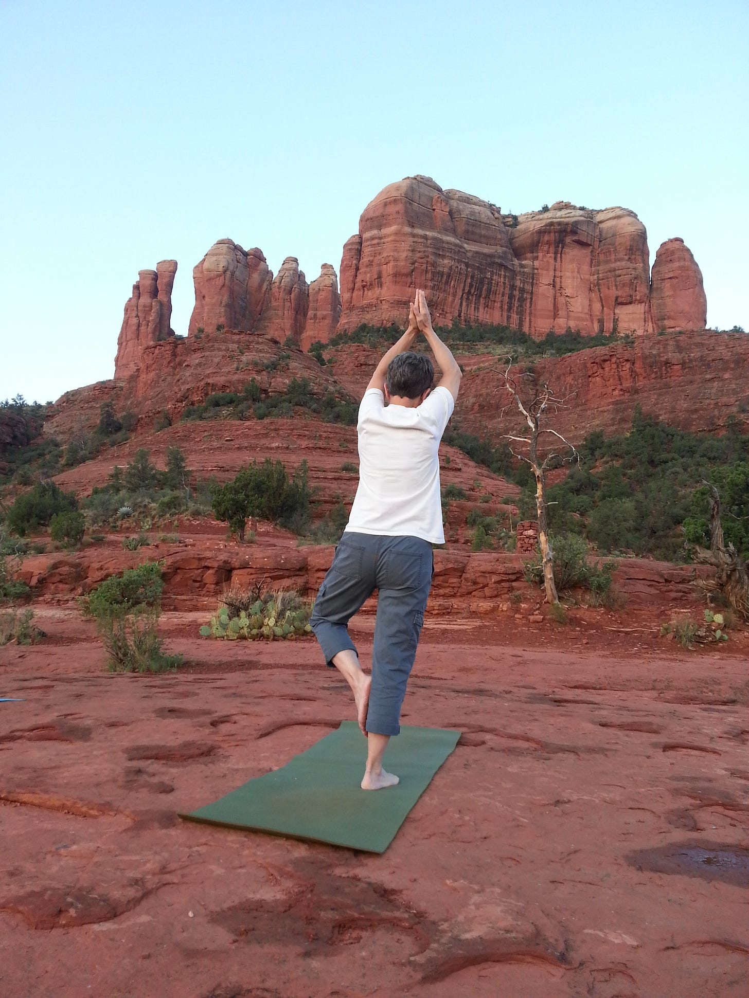 Person standing outside, seen from the back in a yoga tree-pose, facing a tall red rock formation in Sedona at sunrise.