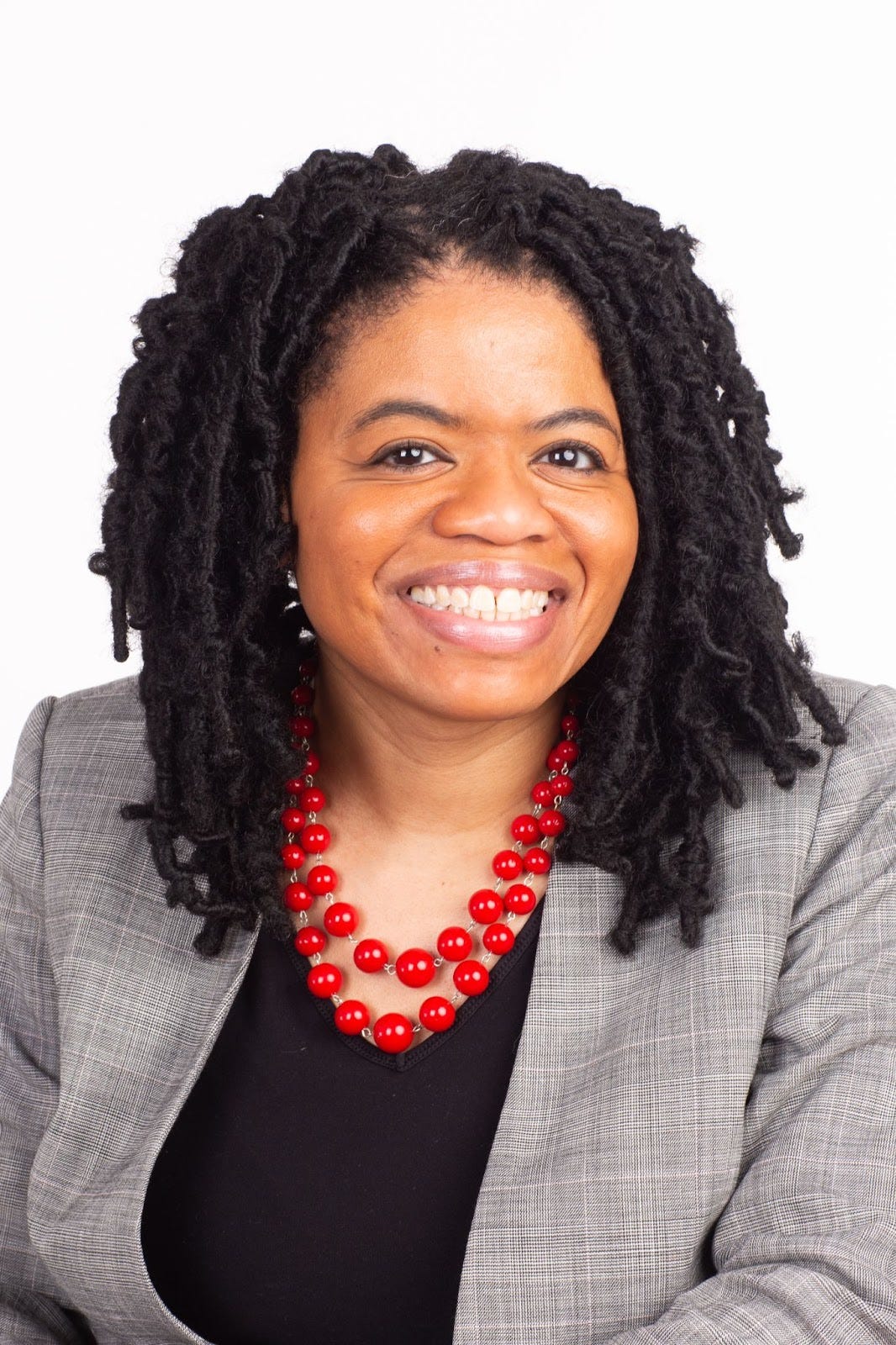 A photo of a smiling Black woman with shoulder-length locs. She is wearing a grey suit jacket and red necklace.