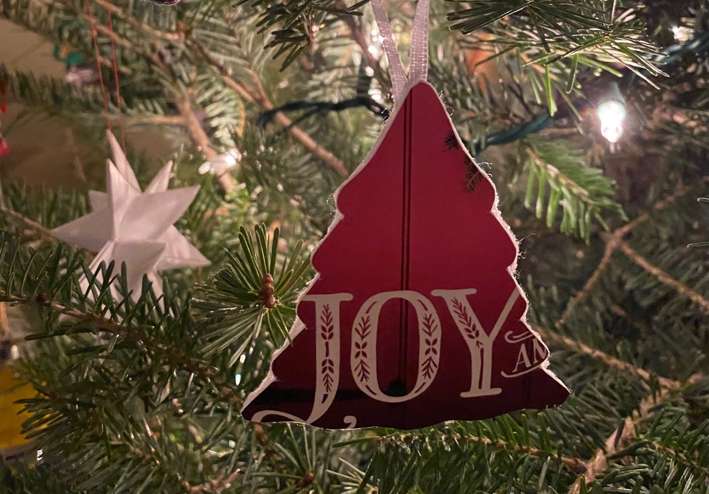 Ornament hanging on tree; paper and cardboard cut out in the shape of a tree with word JOY on it and red barn door in background. Ornament hanging on tree; paper and cardboard cut out in the shape of a tree with word JOY on it and red barn door in background.