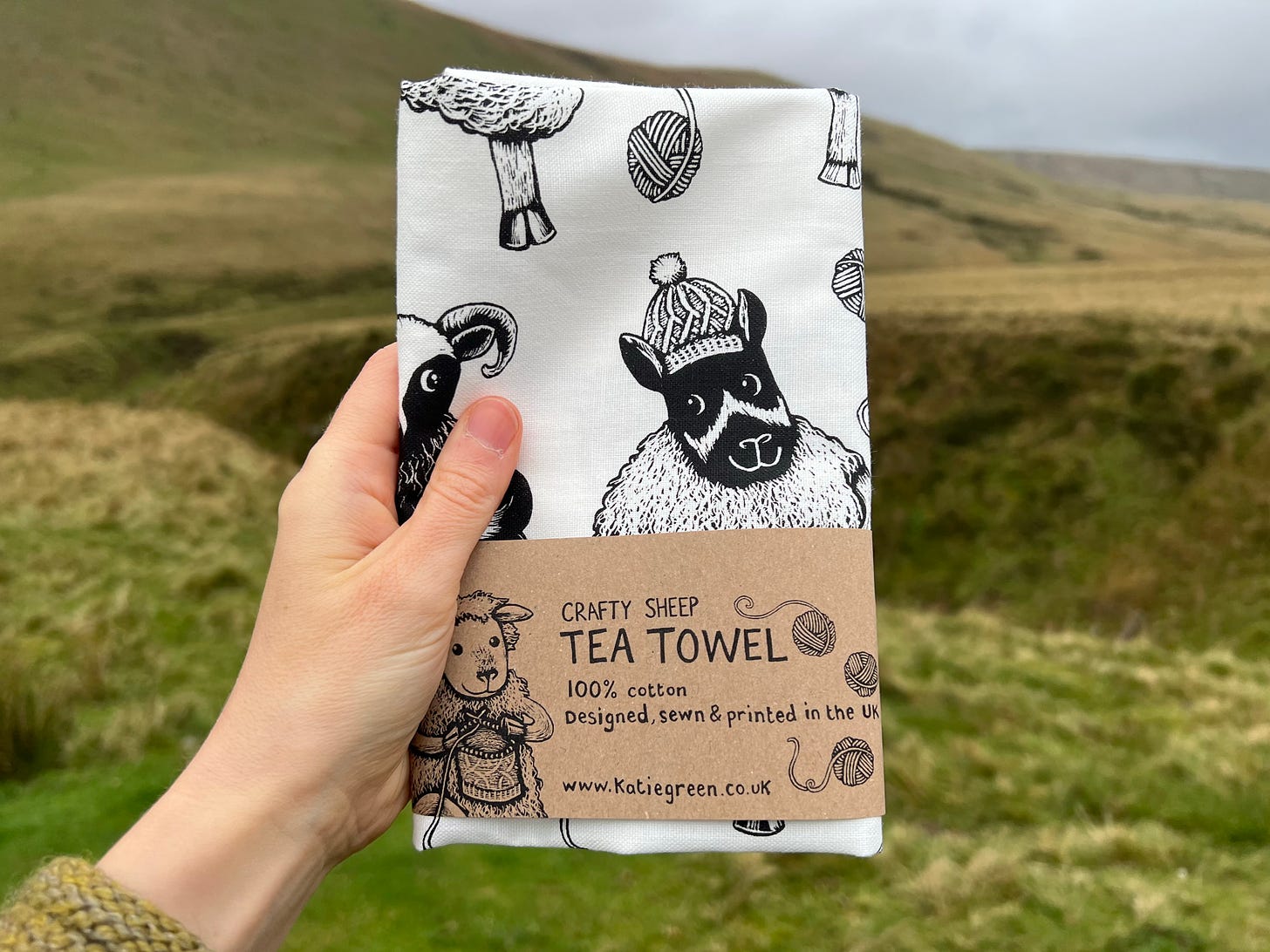 A white tea towel showing craft sheep held up in front of green rolling hills