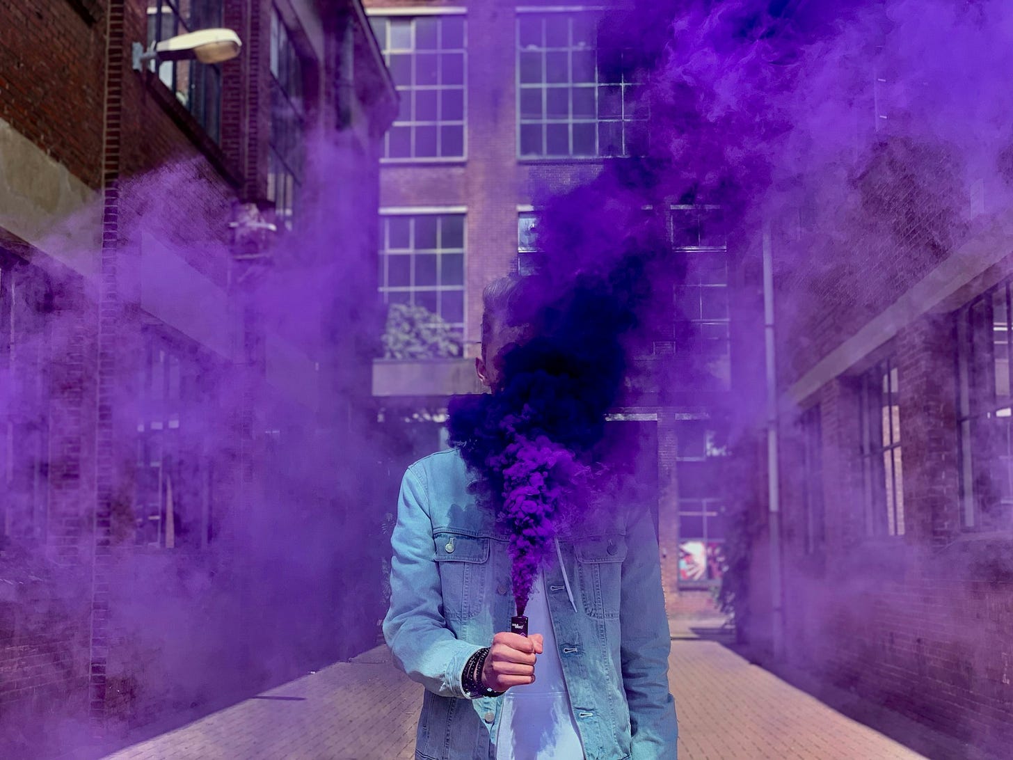 Person stands between two buildings holding a flare whose purple smoke covers their face