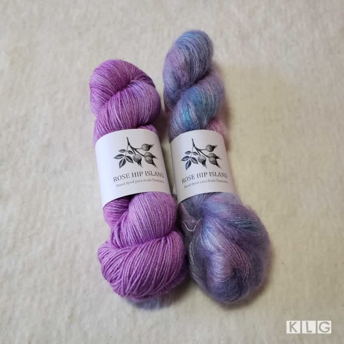 One skein of Rose Hip Island Merino Silk Yarn 4 ply in Purple called Mia Bella. The skein next to it is Mohair Silk Lace called Pixie in blues and purples