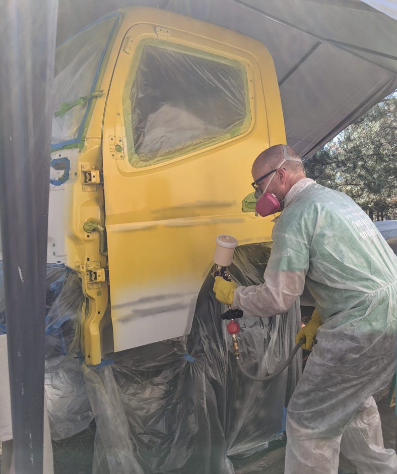 man outside with protective clothing and a respirator mask, painting the white work truck bright yellow with a spray gun