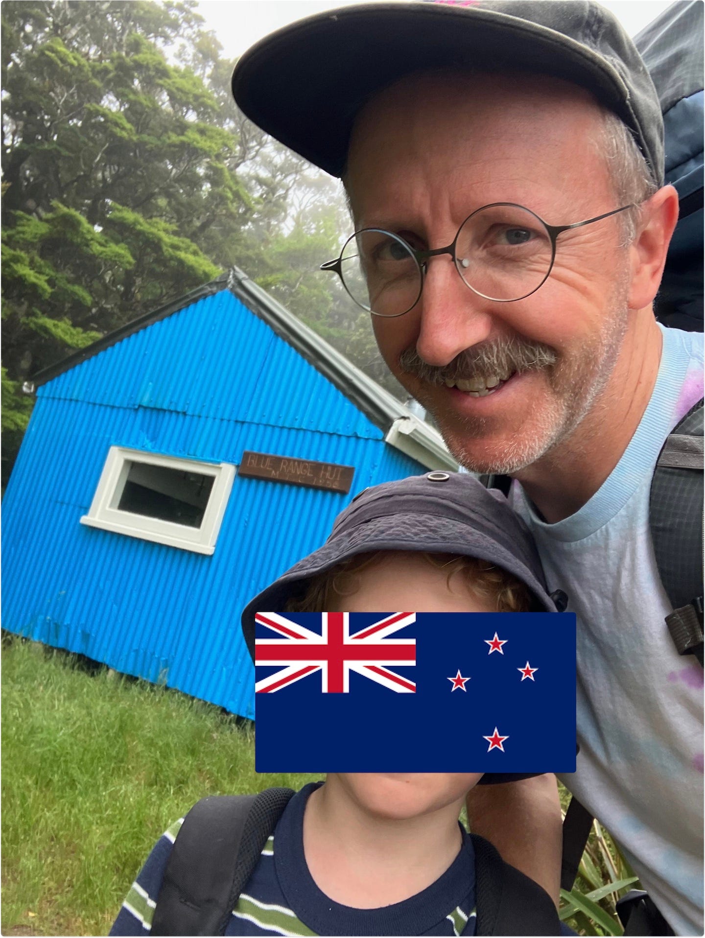 Smiling father with his young son in front of a blue tin hiking hut in a green New Zealand forest.