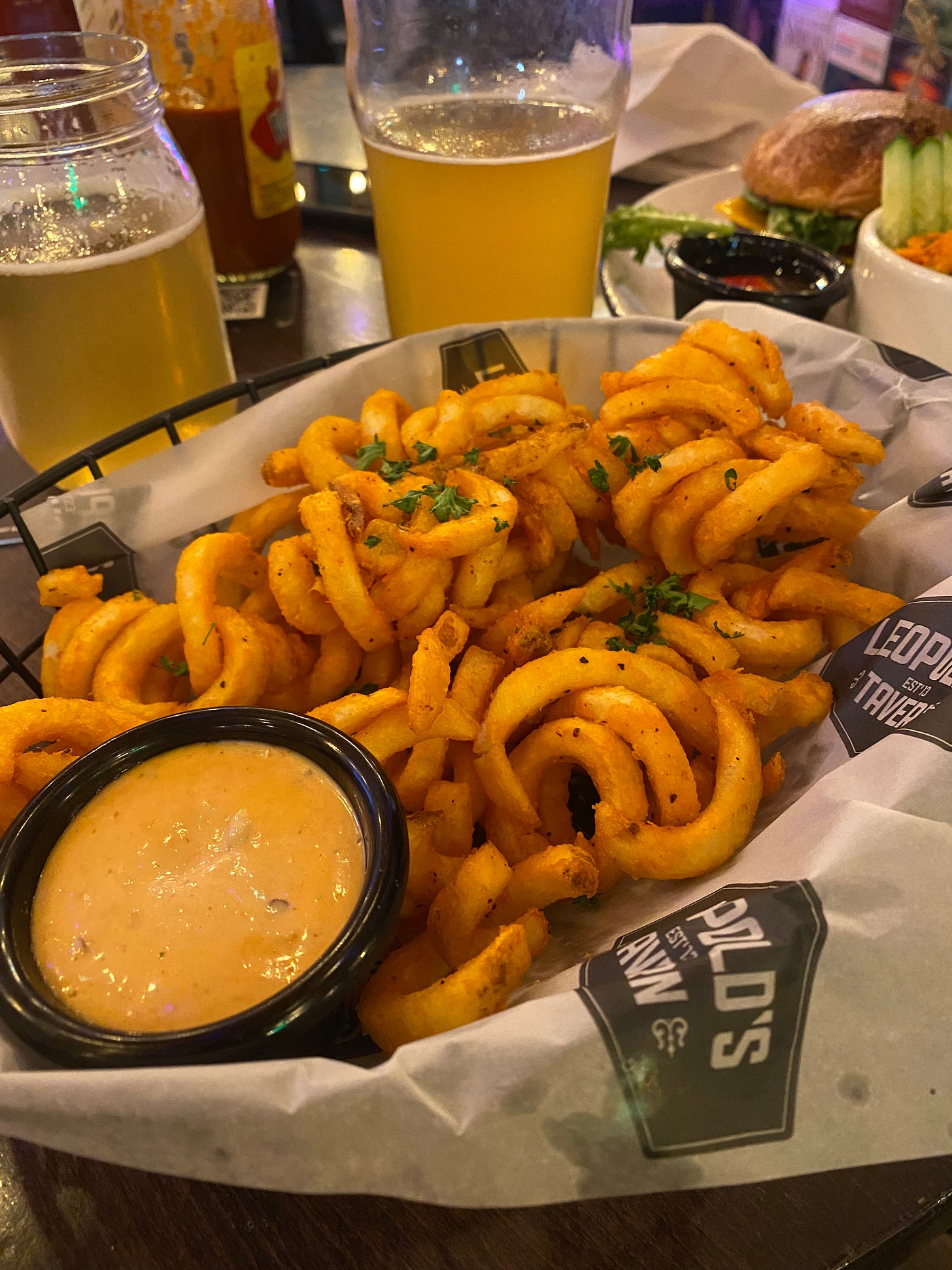 A basket of seasoned curly fries with a small ramekin of chipotle mayo. In the background are two glasses of beer and a bottle of hot sauce.