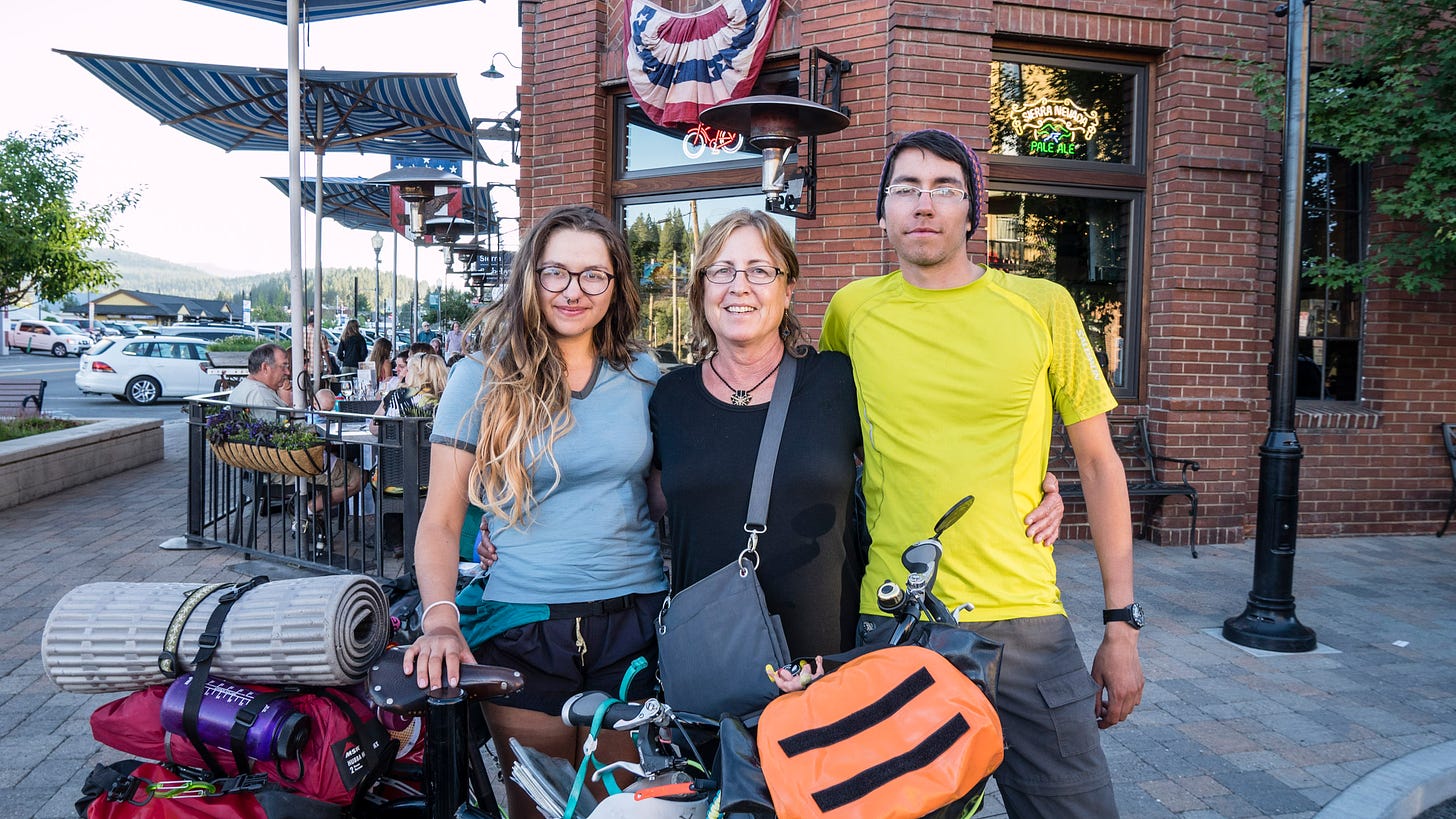 A pair of young bicyclists stand behind a loaded touring bicycle. Between the two siblings is their older aunt. The touring bicycle has bags, water bottles, and a sleeping pad rolled up and bungeed to the back. In the background is the brick facade and entrance of a restaurant with red, white, and blue bunting over the door. Tables of people are sitting outside the restaurant, eating.