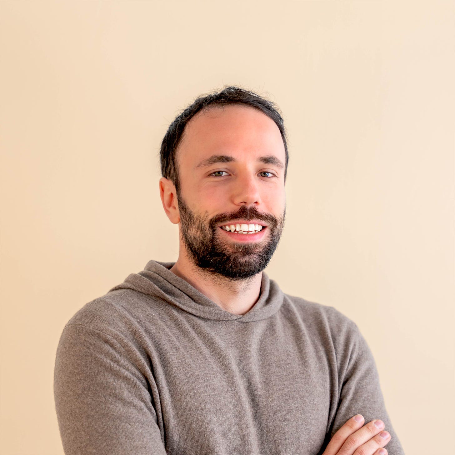 Headshot of Nick, who is a white man with short dark brown hair and a dark brown beard, who is smiling into the camera, wearing a thin grey hoodie, his arms crossed.