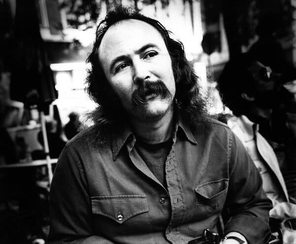 David Crosby, with long (but thinning) hair and a long mustache, seated and gazing into the distance.