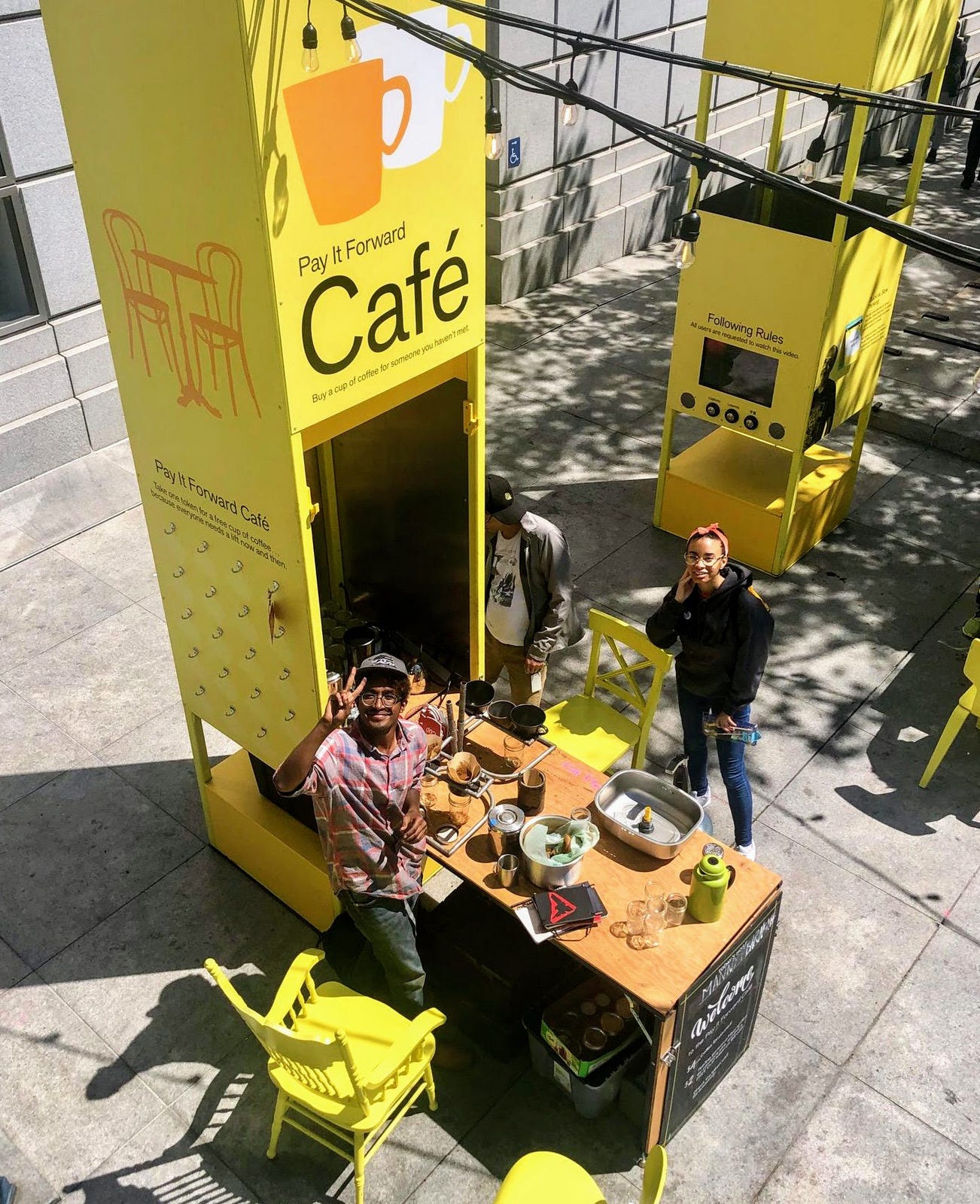 In a shot taken from above, two people stand in a public plaza at a table covered in coffee-making supplies. A tall yellow cabinet Identifies the table as the “Pay it forward café”. Both people are looking up and smiling at the camera.