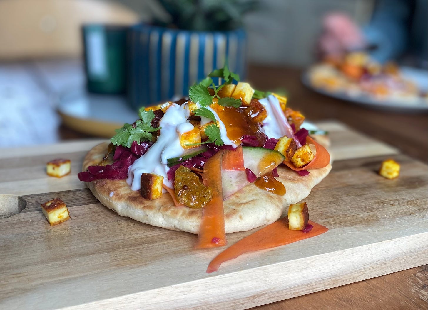 Wooden board with a naan bread topped with carrot ribbons, cucumber, cubed paneer, yoghurt and coriander leaves