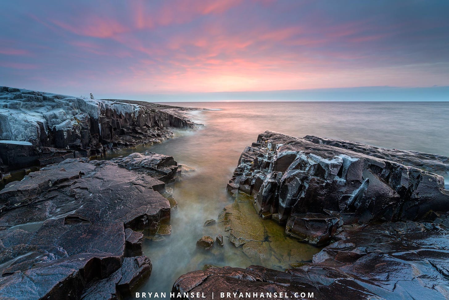 A photo of sunrise over Lake Superior. A basalt shoreline has a crack/slot cut right through it and it aims at a purple and pink sunrise.