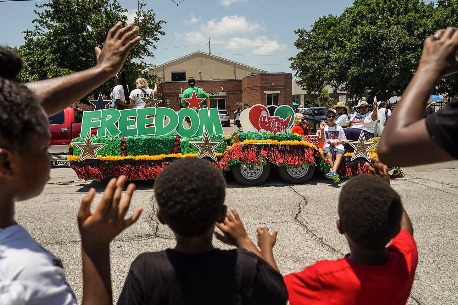 A photo of kids watching a Juneteenth parade. There is one float going by in the photo, which features green, red, and yellow decorations and says “FREEDOM” in big letters.
