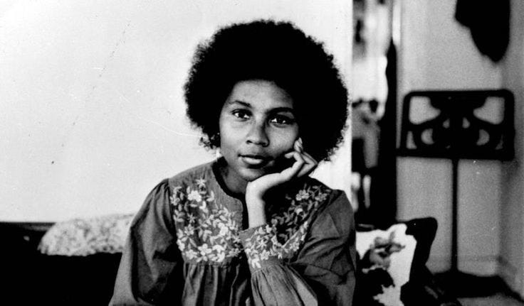 A black-and-white portrait of a young woman with a natural afro hairstyle, resting her chin on her hand, wearing a blouse with embroidered floral patterns. this is bell hooks