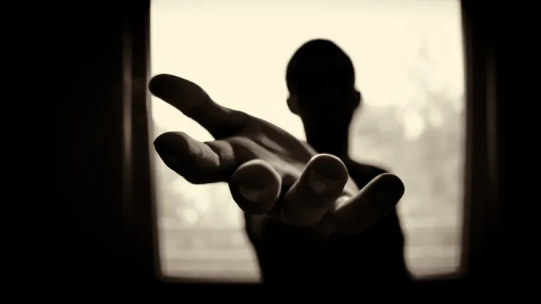 Eine Person im dunklen Zimmer vor einem Fenster streckt die Hand aus.