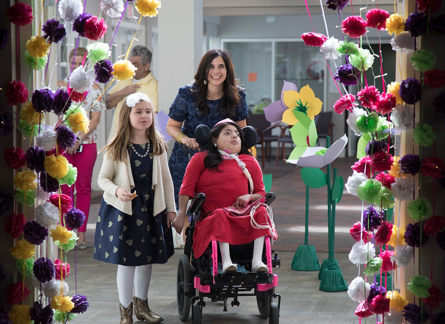 Dalia enters the party surrounded by a floral arch. She holds the hand of a friend. her mother stands behind, pushing her wheelchair and smiles at the camera