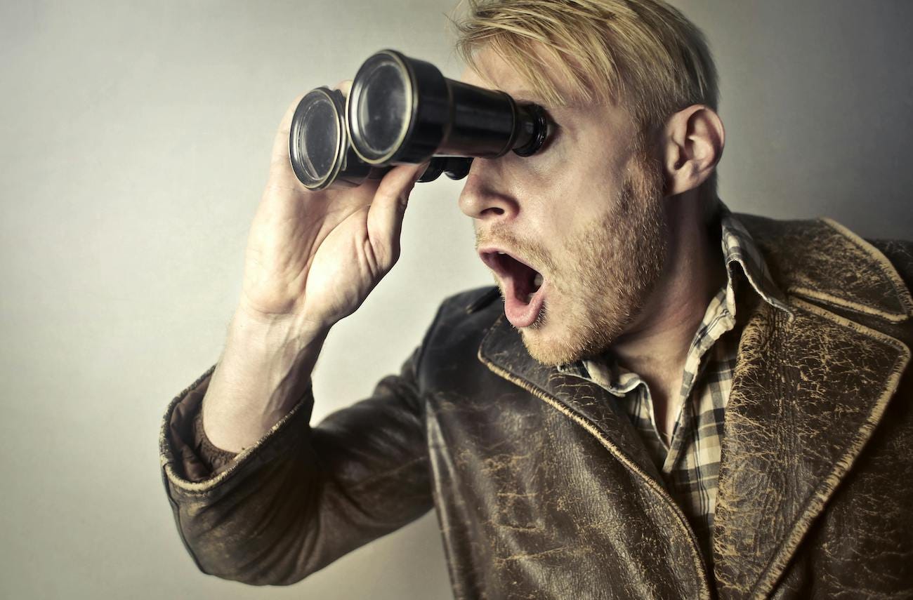 Photo of a man looking through a pair of binoculars with a surprised look on his face. There is no other context in the photo, so we have no idea what he is looking at or why he finds it so shocking.