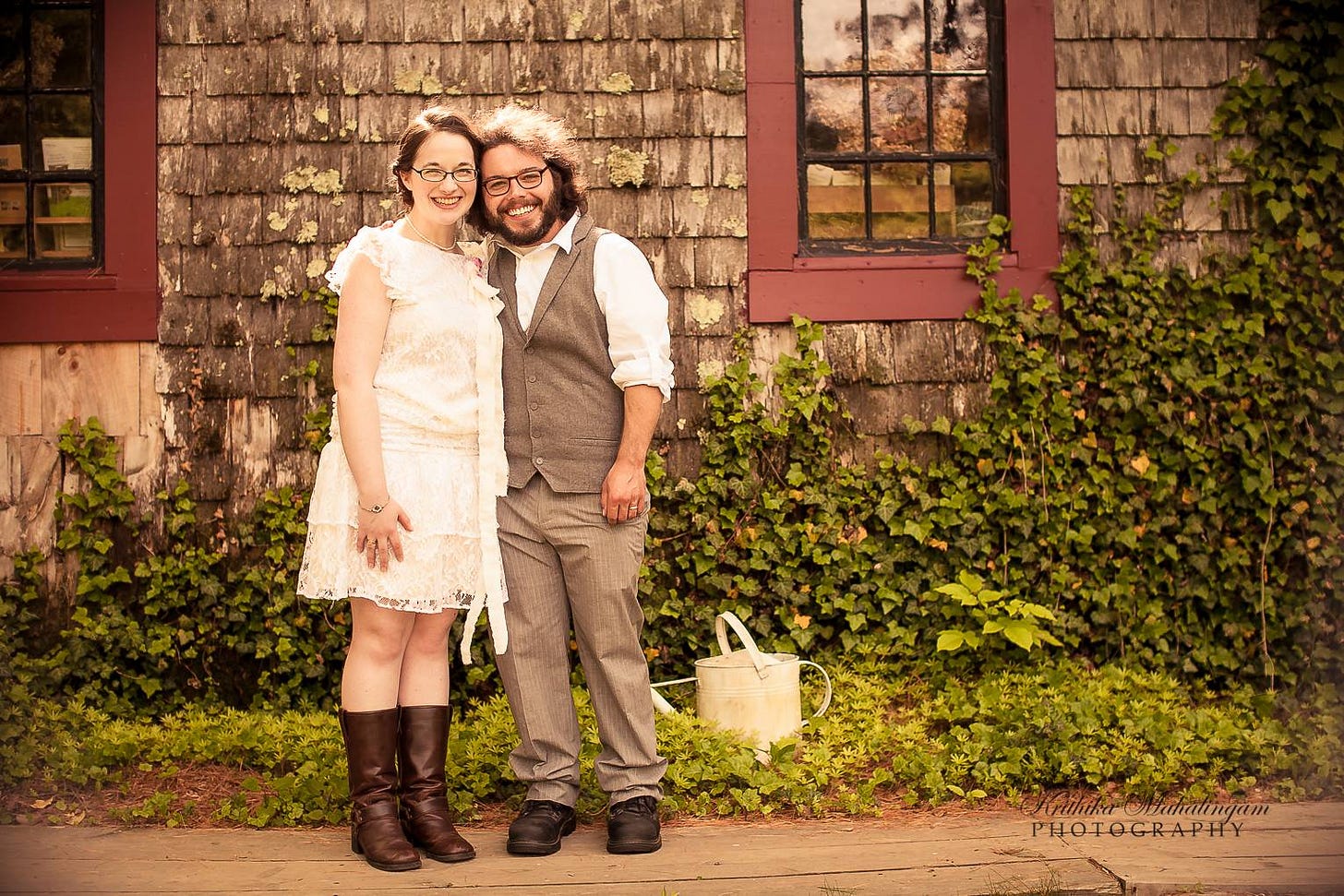 A couple stands in front of a garden space and cedar shank home in their wedding attire, smiling at the camera