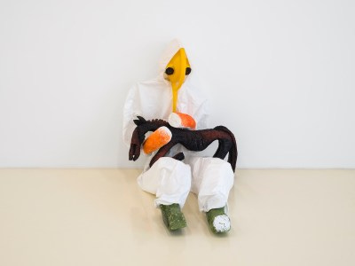 A sculpture of a seated person in a hazmat suit cradling a thin horse. The person runs its hands along the horse's neck. Instead of a face, the person has a yellow object with two black circles for eyes.
