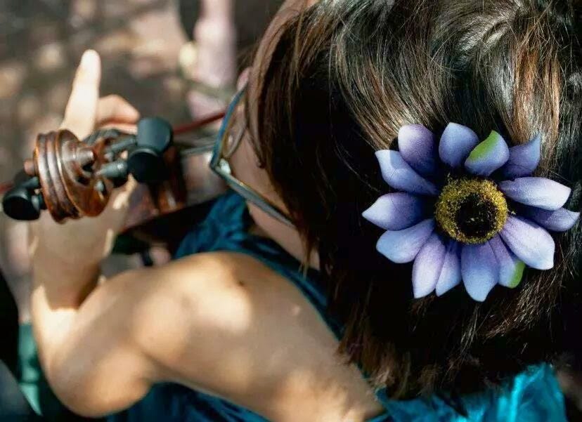 A picture taken from above of Gaelynn playing her violin like a tiny cello. Gaelynn has a purple flower hair-clip in her dark brown hair. The flower has a yellow center and the purple petals are furled out almost like rays of the sun.
