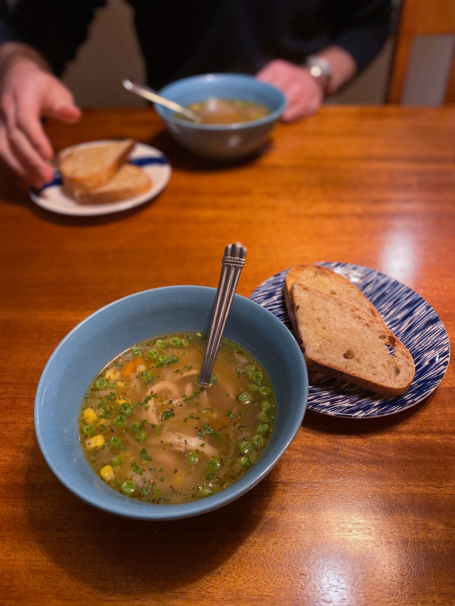 Two blue bowls of chicken noodle soup across from each other on the table, each with a piece of buttered sourdough toast on a small plate beside it. On the opposite side of the table, Jeff's hand reaches for his toast.