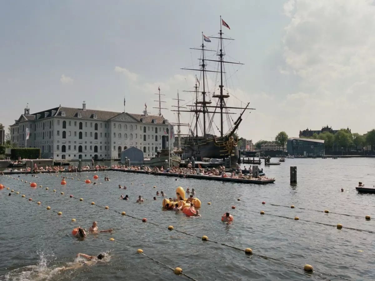 A harbor with a wooden tall ship docked at one pier. In the foreground are swimming lanes market out by buoys. People are clustered on the docks and swimming in the water. There is a large yellow inflatable swan holding several of the swimmers. 